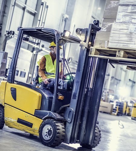 Forklift in foreground moving stack of boxes, warehouse workers in background.